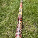 Didgeridoo s barevným ornamentálním vzorem v žlutých, červených a hnědých tónech. Na povrchu je vidět černé kulaté logo s červeným symbolem připomína…