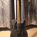 Kytara s matně černým tělem a hnedým hmatníkem, umístěná na stojanu. Nástroj má černý plech a dva humbuckery. Na hlavě kytary je viditelné logo, kter…