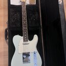Světle modrá elektrická kytara typu Telecaster v černém tvrdém kufru. Na hlavě je viditelné logo Fender. Kytara má bílý pickguard, chromovaný most a…