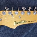 Fotografie zachycuje hlavu dřevěné elektrické kytary v přirozené barvě. Na povrchu je umístěno logo Ibanez a nápisy CUSTOM MADE a ELECTRIC GUITAR. Ná…