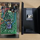 Fotografie zobrazuje vnitřek rozloženého elektronického zařízení. V levé části je kovový šasi s zelenou plošnicí, na které jsou připájková součástka…