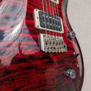 Detail elektrické kytary s červeným plamenovitým povrchem. Nástroj má dva snímače typu humbucker v krémových rámečcích a stříbrný most s fixací strun…