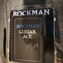 Kytarový procesor v černém obalu, který je umístěn v průhledném plastovém obalu. Na přední straně je bílý a modrý nápis ROCKMAN GUITAR ACE by Tom Sch…