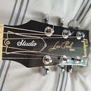 Fotografie zachycuje hlavu černé elektrické kytary. Na povrchu jsou bílé a zlaté nápisy „Studio“, „Les Paul MODEL“ a vertikálně orientované logo „Gib…