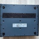 Spodní strana šedého elektronického nástroje s texturovaným povrchem. Uprostřed je černý textilní pás a bílá nálepka s nápisy Arturia MINIBRUTE, Type…