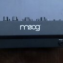 Černý syntetizátor s dřevěnými bočníky. Na přední straně je bílé logo moog®. V dolní části jsou konektory s nápisy AUDIO OUT / ? a 12VDC. Povrch je v…