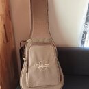 Hnědý polstrovatý pouzdro (gig bag) na kytaru s ramenním popruhem. Na přední kapse je bílý nápis „QUALITY Taylor GUITARS“. Povrch materiálu vypadá či…