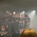 Fotografie zachycuje pět osob na pódiu v koncertním sále. Vlevo stojí muž s akustickou kytarou v rukou, vedle něj stojí čtyři další lidé. V pozadí je…