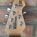 Fotografie zachycuje hlavu kytary z přírodního dřeva s viditnou masivnější strukturou. Na povrchu jsou nápisy MUSIC MAN a Ernie Ball Music Man. V hor…