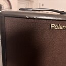 Detail hnědého kytarového komba s černým plastovým rámem a rukojetí. Na přední straně je umístěn kovový nápis Roland. Horní hrana rámu vykazuje známk…