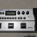 Stříbrný basový efektový procesor s černým pedálem. Na horní části je nápis BOSS GT-1B BASS EFFECTS PROCESSOR a v pravé dolní části nápis COSM. Zaříz…
