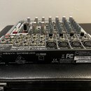 Stříbrný mixážní pult s černými a červenými ovratovými knoflíky. Na zadním panelu je uveden název modelu XENYX QX1202USB, logo behringer a sériové čí…