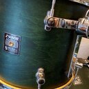 Detailní pohled na tmavě zelený buben s kovovým kováním. Na korpusu je čtvercový štítek s nápisy SONOR, SONIC PLUS II a MADE IN GERMANY. Povrch nástr…