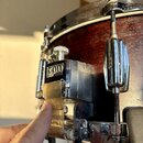 Detailní záběr na chromovaný mechanismus pedálu u červeného dřevěného bubnu. Na kovovém krytu je vidět logo TAMA a pod ním nečitelný nápis. Prst ukaz…