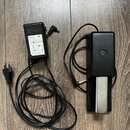 Na fotografii je černý sustain pedál s kovovým šlapadlem a přívodní adaptér. Na horní části pedálu je vyražený nápis Roland. Adaptér má na štítku log…