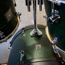 Tři tmavě zelené bubny s kovovým kováním. Na každém z nich je viditelný oválný štítek s nápisy SONOR a SONIC PLUS II MADE IN GERMANY. Povrch nástrojů…