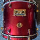 Červený buben s kovovým kováním. Uprostřed korpusu je oválný štít s nápisy „Made Drums“, „TIM“ a nečitelným podpisem pod ním. Povrch nástroje vykazuj…