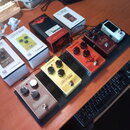 Na černém pedalboardu je umístěno pět efektových pedálů. Zleva doprava jsou vidět pedály s nápisy tc electronic Echobrain ANALOG DELAY (stříbrný), tc…
