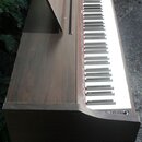 Digitální piano v tmavém dřevěném provedení s bílými a černými klávesami. Na notovém pultu je svisle napsaný nápis GEHANO. Nástroj vykazuje známky op…