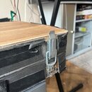 Detailní záběr na hranu a roh černého kufrů z textiledního materiálu s dřevěnou deskou na horní straně. Na boku je vidět kovový zapínací háček. Povrc…