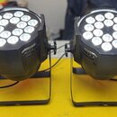 Dvě černé LED par světla na stojáncích. Jsou rozsvícená a každé obsahuje 18 bílých LED diod v kruhovém uspořádání. Povrch je matně černý, bez viditel…