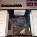 Otevřená krabice s bílým polystyrenem, v níž se nachází modrý USB kabel v plastovém sáčku a uživatelská příručka. Na příručce je viditelné logo AKAI…