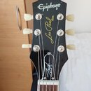 Černá hlava elektrické kytary s bílým logem Epiphone. Uprostřed je zlatý nápis Les Paul MODEL a níže bílý podpis. Na hlavě jsou instalovány šest bílý…