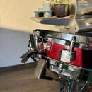 Detailní záběr na červený buben s chromovaným obručím a ladicími šrouby. Na boku je vidět mechanismus pro napnutí blány a černá textilní stužka. Nást…