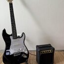 Černá elektrická kytara s bílým pickguardem a světle hnědým hmatníkem, umístěná na černém stojanu. Na hlavě kytary je viditelné logo Fender. Vedle ky…