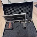 Otevřený hliníkový přepravní kufr s černou vnitřní výplní z akustické pěny a tmavě šedým kobercovým povrchem dna. Uvnitř se nachází černý zdroj s náp…