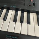Detail klaviatury elektronického nástroje s černým povrchem. Jsou vidět bílé a černé klávesy, které vykazují známky opotřebení a znečištění. Nad kláv…