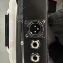 Černý obdélníkový přístroj s kovovými konektory. Na straně jsou vidět jeden XLR vstup/výstup, dva jack konektory a přepínač. Povrch je černý s vidite…