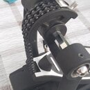 Černý bicí pedál s řetízkovým přenosem. Konstrukce je kombinací černého kovu a stříbrných kovových prvků. Na zařízení nejsou viditelné žádné značky a…