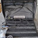 Černý kovový pedalboard s nápisem ROCKBOARD v horní části. Na desce je umístěn napájecí zdroj s nápisem PERA POWER ALUMIC a připojeno několik černých…