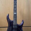 Tmavě fialová elektrická kytara s černým hmatníkem a dvěma humbuckery. Na hlavě je bílý nápis MUSIC MAN. Nástroj stojí na černém stojanu před dřevěno…