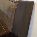 Detailní pohled na pravou boční část hnědého pianina s lakovaným povrchem. Nástroj je umístěn u bílé stěny, na které visí dva obrazy v rámech. V odra…