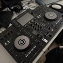 Černý DJ 컨트롤러 s dvěma jogovými koly a centrálním mixážním sekcí. Na levé straně je viditelný nápis XDJ-RR a v pravé části logo Pioneer DJ. Za přístro…