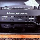Černý elektronický perkusní nástroj s bílým nápisem HANDSonic. Zadní panel obsahuje USB porty označené HI-SPEED USB MEMORY a COMPUTER, MIDI konektory…