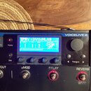 Černý efektový procesor s nápisem VOICELIVE 2. Modrý displej zobrazuje text ZPEV + DVOJHLAS, hodnoty PRESET 1, STEP 1, COUNT 2 a SHORTCUT CHOIR OFF.…