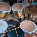 Fotografie zobrazuje bicí sadu s mosaznými nebo zlatými kotli dvou tomů a dřevěným kotlem jednoho tahu. Sada obsahuje dva tomy, snare drum s bílýmI s…