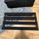 Na fotografii je černý pedalboard s čtyřmi paralelními lištami. V horní části je umístněn černý přepravní vak s bílým nápisem ROCKBOARD. Povrch pedal…