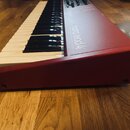 Červený syntetizátor s klávesnicí položený na dřevěné podlaze. Na červeném povrchu je bílý nápis nord lead 4. Nástroj vykazuje mírné opotřebení v pod…