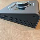 Šedý kovový audio interface s velkým ovládacím knoflíkem na horní straně. Na pravém horním rohu je nápis apollo twin. Boční stěna zařízení obsahuje s…