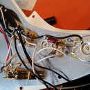 Fotografie zachycuje vnitřní elektroinstalaci elektrické kytary s bílým pickguardem a stříbrnou stínovkou. Jsou vidět tři snímače, potenciometry a za…