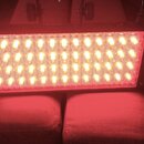 Obdélníkový LED panel s černým rámem a chladicími žebry po stranách. Panel je rozsvícen žlutobílým světlem a vyzařuje červený odlesk. Na horní hraně…