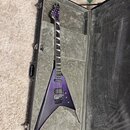 Fialová elektrická kytara s ostře hranatým tělem typu V v tvrdém kufru. Na hlavě hmatníku je vidět bílé logo Jackson. Kytara má dva humbuckery, Floyd…
