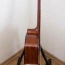Kytara akustického typu v hnědé barvě s lakovaným povrchem. Nástroj je umístěn na černém stojanu v rohu místnosti. Na fotce nejsou viditelné žádné zn…