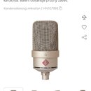 Snímek zobrazuje kondenzátorový mikrofon stříbrné barvy s kovovou síťovkou. Na těle je viditelné červené kosočtvercové logo a nápis NEUMANN MICROPHON…