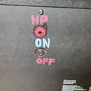 Zadní strana černého komba s texturovaným povrchem. U vstupů jsou ručně nanesené nápisy červenou a bílou barvou: „HP“, „ON“ a „OFF“. V pravém dolním…