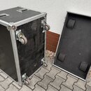 Černý transportní case s hliníkovými hranami a rohovými zpevněními. Case je vybaven čtyřmi kolečky a dvěma madly na horní straně. Vedle stojí otevřen…